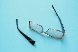 Old broken eyeglasses with damaged legs on blue background. Poor eyesight. Reuse and repair concept. Idea of health. Failure optic eyewear. Breakage of vision correction glasses. Close up, flat lay photo