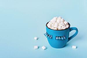 Blue monday concept. Blue mug of cocoa with marshmallows on blue background. Mug is decorated with eyelashes down photo