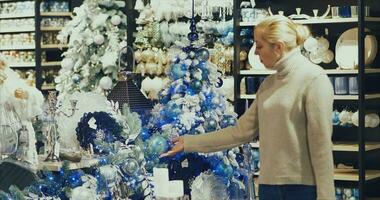 ein blond Frau Spaziergänge durch ein groß Einkaufen Center gegen das Hintergrund von dekoriert Weihnachten Bäume. suchen beim ein Anzeige Fall mit Neu Jahre Dekorationen. Neu video