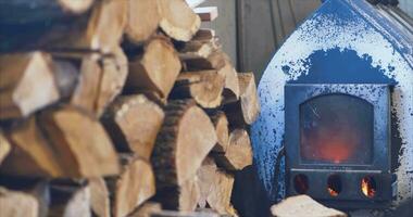 A fire burns in a metal wood-burning stove against the background of stacked firewood in the carpentry workshop. Close-up. slow motion. CINEMATIC video