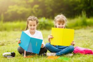 two cute multicultural schoolgirls sitting on lawn under tree and reading book together photo