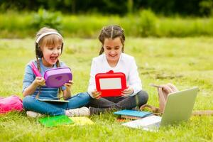 Pupils of primary school with lunch-boxes in hands. Girls with backpacks are eating on the lawn. Beginning of lessons. First day of fall. photo