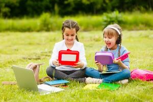 Pupils of primary school with lunch-boxes in hands. Girls with backpacks are eating on the lawn. Beginning of lessons. First day of fall. photo