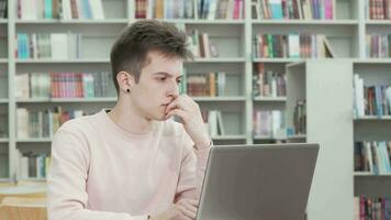 Jeune homme à la recherche réfléchi tandis que travail sur une portable à le bibliothèque video