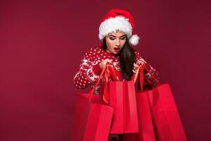 Young pretty woman or girl with long beautiful hair in Santa hat and New Year's dress holding red package on red studio background photo