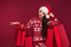 joven bonito mujer o niña con largo hermosa pelo en Papa Noel sombrero y nuevo años vestir participación rojo paquete en rojo estudio antecedentes foto