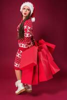 Young pretty woman or girl with long beautiful hair in Santa hat and New Year's dress holding red package on red studio background. Full-length photo