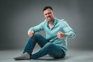 The man is sitting on the floor, isolated on gray background. Man showing different emotions. photo