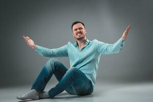 Handsome young man sitting on a floor with raised hands, isolated on gray background photo