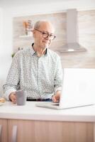 Grayhaired senior retired man browsing on internet. Daily life of senior man in kitchen during breakfast using laptop holding a cup of coffee. Elderly retired person working from home, telecommuting using remote internet job online communication photo