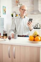Retired old man watching video watching video on smartphone in kitchen while having breakfast. Using modern technology while enjoying morning coffee during breakfast. Authentic portrait of retired senior internet online technology photo