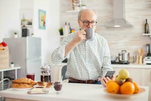 mayor hombre Bebiendo café durante desayuno en ktichen y utilizando teléfono inteligente foto