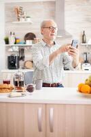 Mature man surfing the social media using phone during breakfast in kitchen. Elderly person using internet online chat technology video webcam making a video call connection camera communication conference call photo