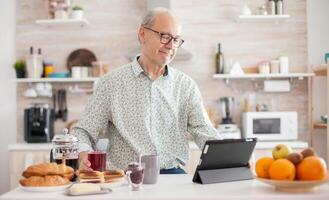 Senior person browsing on tablet pc in kitchen during breakfast having a healthy lifestyle. Elderly person with tablet portable pad PC in retirement age using mobile apps, modern internet online information technology with touchscreen photo