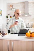 Elderly man surfing on internet using tablet computer during breakfast in kitchen. Senior person with tablet portable pad PC in retirement age using mobile apps, modern internet online information technology with touchscreen photo