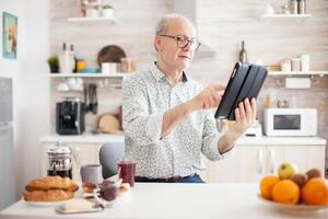 Happy elderly person with portable tablet PC in kitchen during breakfast. Senior person with tablet portable pad PC in retirement age using mobile apps, modern internet online information technology with touchscreen photo