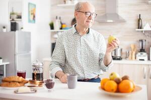 Mature man holding green apple before taking a bite during breakfast in kitchen. Elderly retired person eating apple during breakfast in modern cozy kitchen. Healthy green happy fresh lifestyle photo