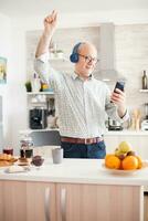 Happy senior man listening music wearing headphones in kitchen during breakfast. Elderly retired person enjoying modern fun happy lifestyle, dancing relaxed, smiling and using technology photo