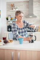 mayor mujer prensado en francés prensa a hacer sabroso café durante desayuno en cocina. mayor persona en el Mañana disfrutando Fresco marrón café Café exprés taza cafeína desde Clásico taza, filtrar relajarse refresco foto