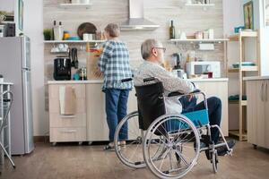 Pensive disabled elderly person in wheelchair looking on the window from the kitchen. Disabled man sitting in wheelchair in kitchen looking through window while wife is preparing breakfast. Invalid, pensioner, handicapped, paralysis. photo
