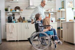 Senior man taking grocery paper bag from handicapped wife in wheelchair. Mature people with fresh vegetables from market. Living with disabled person with walking disabilities photo