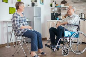 Old woman and her disabled husband in wheelchair chatting in kitchen.Elderly person having a conversation with husband in kitchen. Living with disabled person with walking disabilities photo