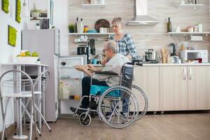 Handicapped senior man in wheelchair taking eggs carton from refrigerator for wife in kitchen. Senior woman helping disabled husband. Living with disabled person with walking disabilities photo