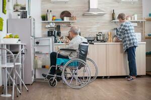 Disabled senior man in wheelchair taking eggs carton from refrigerator for wife in kitchen. Senior woman helping handicapped husband. Living with disabled person with walking disabilities photo