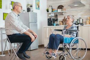 Retired invalid woman in wheelchair having a conversation with old elderly husband in kitchen. Old man talking with wife. Living with disabled person with walking disabilities photo