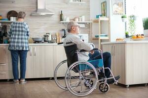 Disabled man sitting in wheelchair in kitchen looking through window while wife is preparing breakfast. Invalid, pensioner, handicapped, paralysis. photo