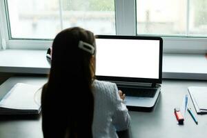 Smart Little Girl Does Homework in Her Living Room. She's Sitting at Her Desk Writes with a Pen in Her Textbooks and Uses Laptop. photo