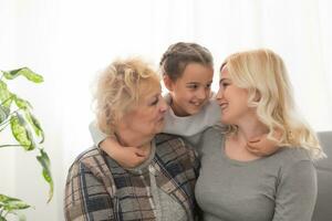 Three generations of women. Beautiful woman and teenage girl are kissing their granny while sitting on couch at home photo