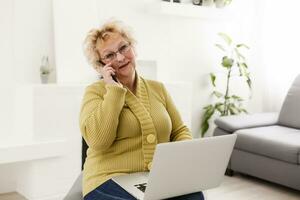 Attractive elderly woman sitting on couch at home, looking at camera, makes video call, communication distantly, modern wireless technologies concept photo