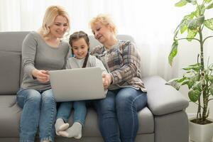 Happy three generations of women sit relax on couch laugh watching funny video on laptop, smiling positive females grandmother, mother and daughter have fun rest on sofa enjoy movie on computer photo