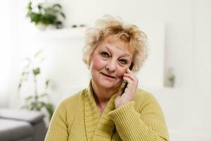 Attractive elderly woman sitting on couch at home, looking at camera, makes video call, communication distantly, modern wireless technologies concept photo