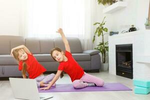 dos niñas practicando yoga, estiramientos, fitness por video en el cuaderno. entrenamiento de educación en línea distante, aeróbico en casa. estilo de vida saludable, coronavirus, quédate en casa. los niños hacen deporte en casa en cuarentena. foto