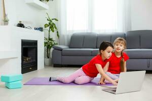 two little girls practicing yoga, stretching, fitness by video on notebook. Distant online education training, aerobic at home. Healthy lifestyle, coronavirus, stay home. Kids sport home quarantine. photo