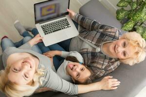 Happy three generations of women sit relax on couch laugh watching funny video on laptop, smiling positive females grandmother, mother and daughter have fun rest on sofa enjoy movie on computer photo