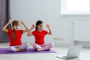 two little girls practicing yoga, stretching, fitness by video on notebook. Distant online education training, aerobic at home. Healthy lifestyle, coronavirus, stay home. Kids sport home quarantine. photo
