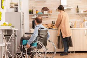 Man with walking handicap in wheelchair looking at wife how she's cooking. Disabled paralyzed handicapped man with walking disability integrating after an accident. photo