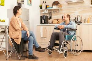 Handicapped man in wheelchair looking at caring wife in kitchen. Disabled paralyzed handicapped man with walking disability integrating after an accident. photo