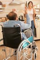 Wife screaming in kitchen to her disabled man in wheelchair while having a disagreement. Disabled paralyzed handicapped man with walking disability integrating after an accident. photo