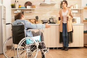 Wife looking angry at disabled man in wheelchair during an argument about their relationship in kitchen. Disabled paralyzed handicapped man with walking disability integrating after an accident. photo