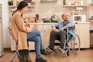 Wife crying because of disagreement with disabled husband in wheelchair. Couple argue in kitchen. Disabled paralyzed handicapped man with walking disability integrating after an accident. photo