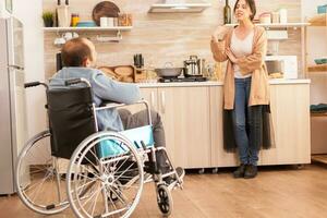 Unhappy man in wheelchair having a disagreement with wife in kitchen. Woman shouting at handicapped man. Disabled paralyzed handicapped man with walking disability integrating after an accident. photo