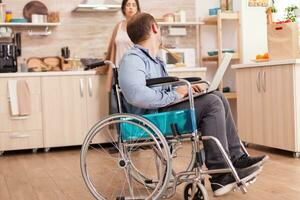 Disabled man in wheelchair working on laptop while wife is cooking in kitchen. Disabled paralyzed handicapped man with walking disability integrating after an accident. photo