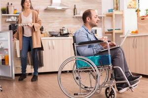 Disabled man in wheelchair during video call on laptop in kitchen while is holding box of eggs and holding refrigerator door open. Disabled paralyzed handicapped man with walking disability integrating after an accident. photo