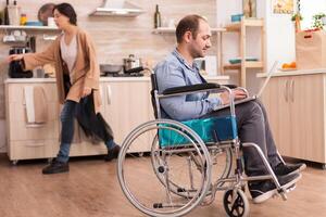 Invalid man in wheelchair working remotely on laptop in kitchen while is standing in front of refrigerator. Disabled paralyzed handicapped man with walking disability integrating after an accident. photo