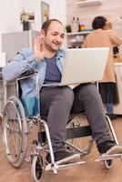 Disabled entrepreneur in wheelchair waving during a video call on laptop while wife is cooking lunch. Disabled paralyzed handicapped man with walking disability integrating after an accident. photo