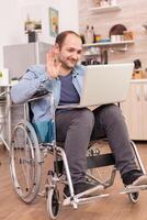 empresario en silla de ruedas ondulación durante un vídeo conferencia en ordenador portátil en cocina mientras es preparando alimento. discapacitado paralizado minusválido hombre con caminando invalidez integrando después un accidente. foto